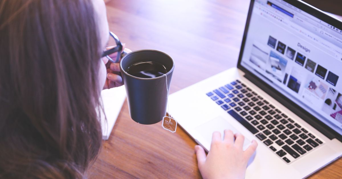 A woman enjoys coffee while browsing the web on a laptop at a home office desk.